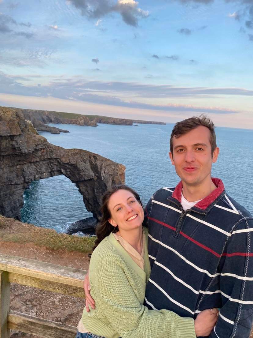 Joe and Naomi stood in front of the green bridge of Wales, with the natural rock arch behind them, surrounded by the sea.