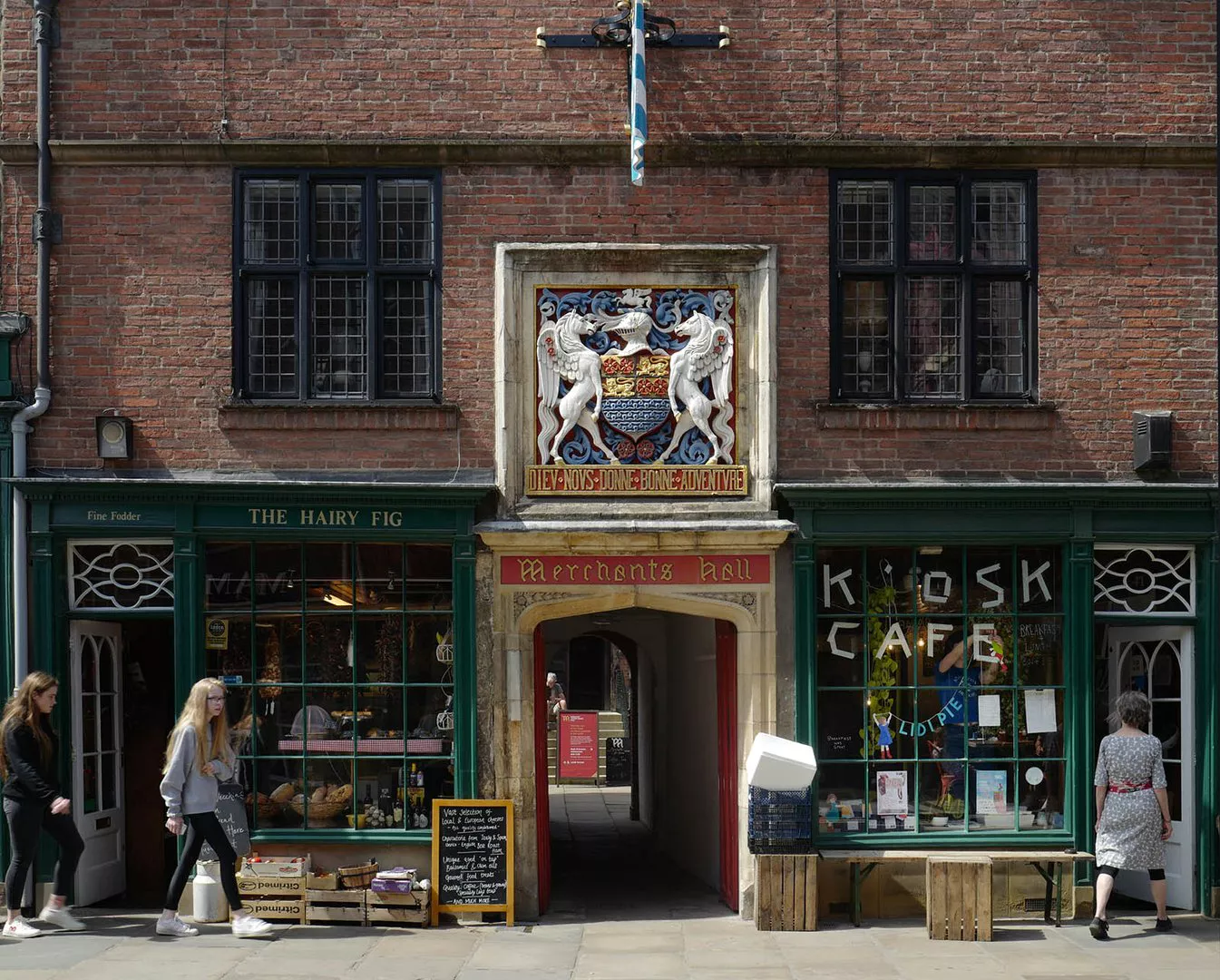 Photo of the entrance to the hall from Fossgate. The entrance is a covered passage, with Merchants Hall written over it along with a coat of arms, with two shops either side of the passage. Notably on the right is Kiosk Cafe.